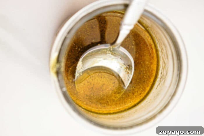 Effortless Slow Cooker Vegetables 5 Close-up view of a glass jar filled with a golden-brown liquid, likely a sauce or dressing perfect for slow cooker vegetables, with a metal spoon partially submerged in it. The liquid has some visible seasoning or spices. The background is white and minimal.