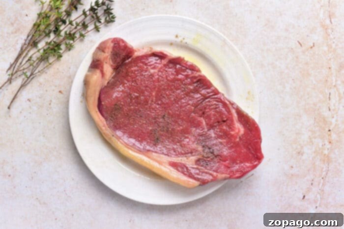 Cooking Beef Strip Loin Steaks to Perfection 3 A raw, beautifully marbled beef strip loin steak seasoned with a generous sprinkle of coarse salt and black pepper, accompanied by fresh sprigs of thyme. This preparation highlights the simple yet effective seasoning before cooking, showcasing a crucial step for achieving perfect pan-seared beef strip loin steaks.