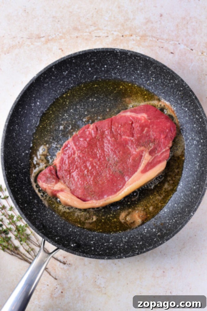 Cooking Beef Strip Loin Steaks to Perfection 4 A raw, seasoned strip loin steak, ready for cooking, sits in a hot frying pan with a small amount of oil, showcasing the initial stages of pan-searing. A bundle of fresh herbs, likely thyme, rests beside the pan on a light-colored, textured surface, indicating the aromatic additions used during the cooking process to enhance flavor.