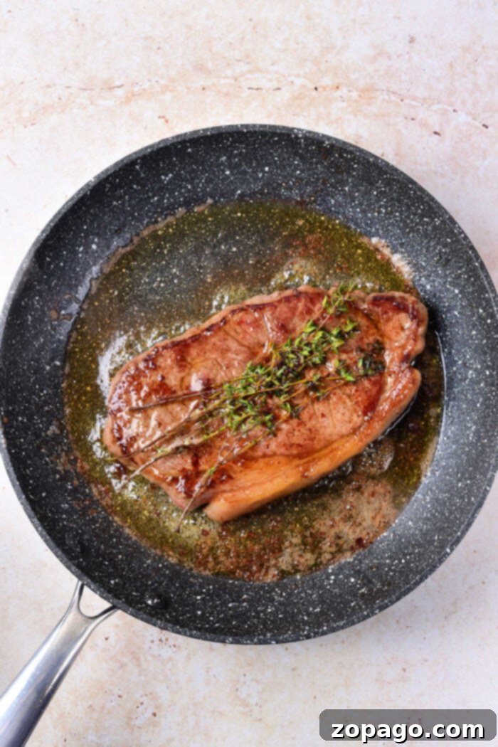 Cooking Beef Strip Loin Steaks to Perfection 5 A perfectly cooked strip loin steak, garnished with fresh thyme sprigs, resting in a black non-stick frying pan. The steak displays a beautiful, even sear and a glistening, juicy surface, highlighting the successful pan-searing technique. The melted fat and pan juices create a small pool around the steak, inviting a delicious tasting experience of expertly cooked beef strip loin steaks.
