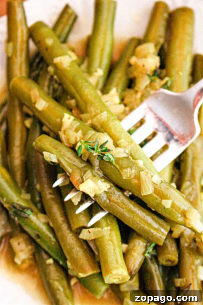 Effortless Crockpot Green Beans 2 A close-up image of crockpot-cooked green beans seasoned with chopped onions and herbs. A fork is piercing several of the green beans, and the dish appears to be served on a white plate.