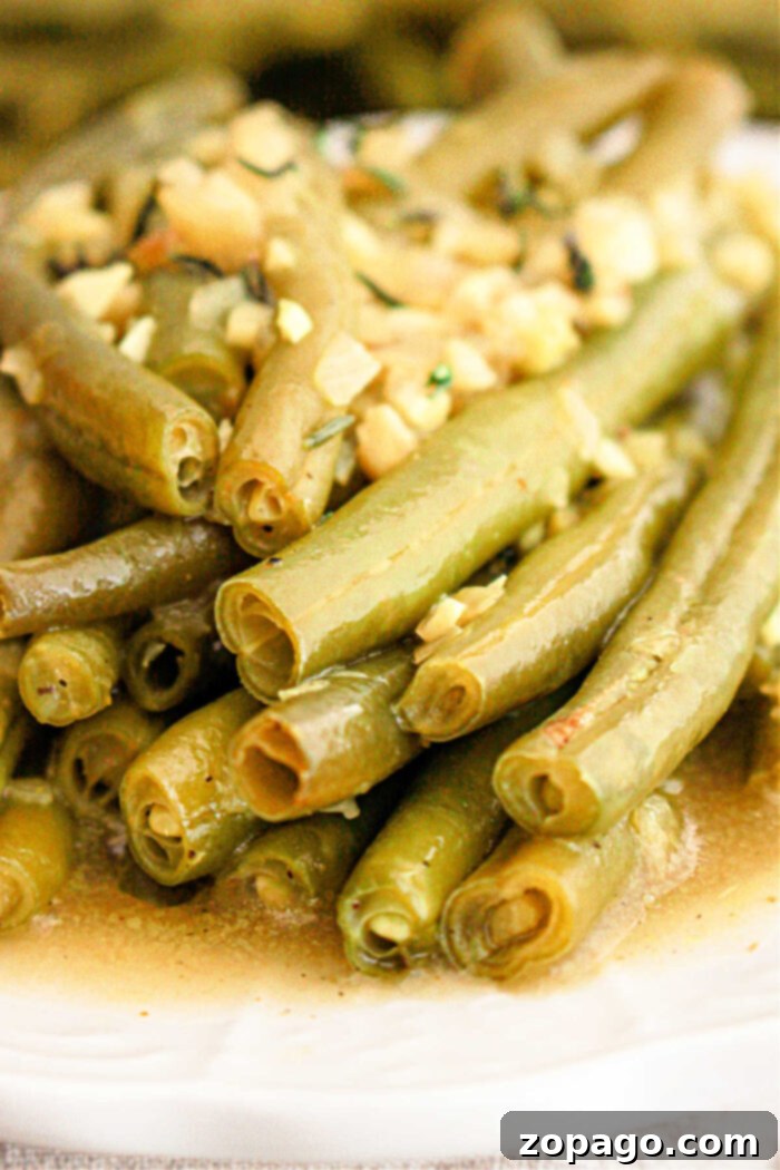 Effortless Crockpot Green Beans 6 A close-up of crockpot-cooked green beans topped with finely chopped garlic and herbs. The green beans are slightly glossy from the seasoning and arranged in a pile on a white plate.