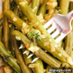 Effortless Crockpot Green Beans 7 A close-up image of crockpot-cooked green beans seasoned with chopped onions and herbs. A fork is piercing several of the green beans, and the dish appears to be served on a white plate.