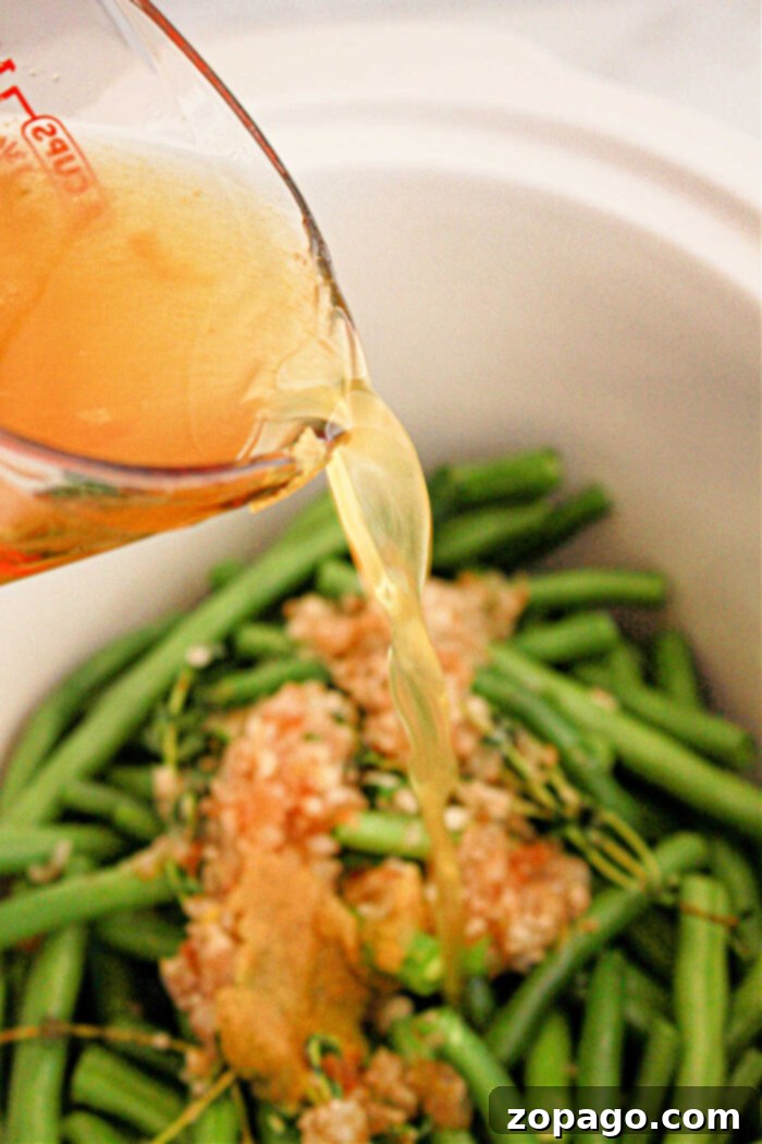 Effortless Crockpot Green Beans 8 Close-up of a measuring cup pouring broth into a crockpot filled with green beans, minced garlic, and other seasonings. The green beans are uncooked, and the garlic appears to be spread on top.