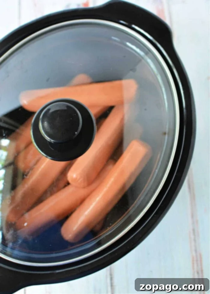 Rapid Slow Cooker Hot Dogs 2 A black slow cooker with a glass lid contains several slow cooker hot dogs heating inside. The slow cooker is placed on a white surface with a faint pattern.