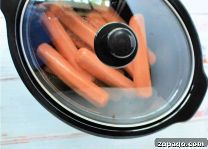 Rapid Slow Cooker Hot Dogs 5 A black slow cooker with several slow cooker hot dogs inside, viewed from the top. The glass lid partially obscures the hot dogs. The slow cooker rests on a light-colored surface.