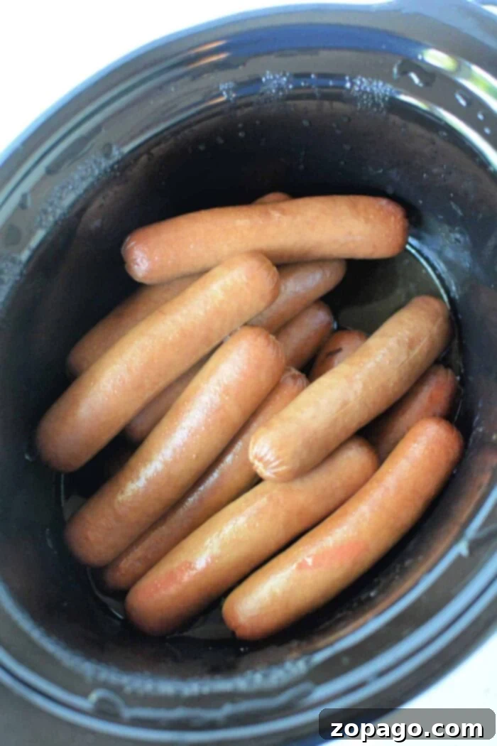 Rapid Slow Cooker Hot Dogs 6 A black slow cooker filled with neatly arranged Slow Cooker Hot Dogs. The hot dogs are light brown and have a glossy appearance, indicating they have been recently cooked.