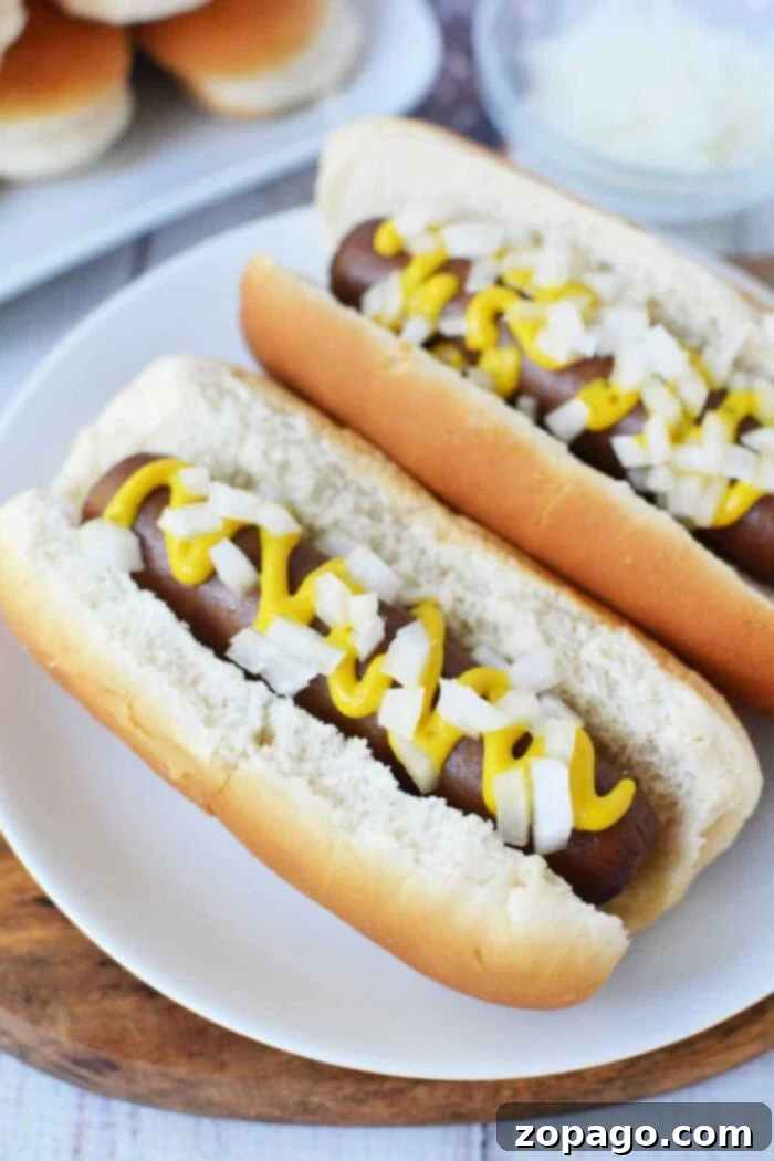 Rapid Slow Cooker Hot Dogs 7 Two hot dogs in buns are placed on a white plate. The hot dogs, slow-cooked to perfection, are topped with diced onions and drizzled with yellow mustard. A platter of additional slow cooker hot dogs and a bowl of chopped onions are visible in the background.