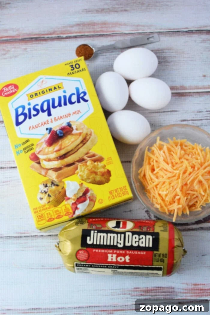 Savory Sausage Biscuits 3 A vibrant flat lay of ingredients for savory muffins: a classic Bisquick box, three fresh eggs, a mound of shredded cheddar cheese, a package of hot Jimmy Dean sausage, and a small spoonful of cayenne pepper, all neatly arranged on a pristine white wooden surface.