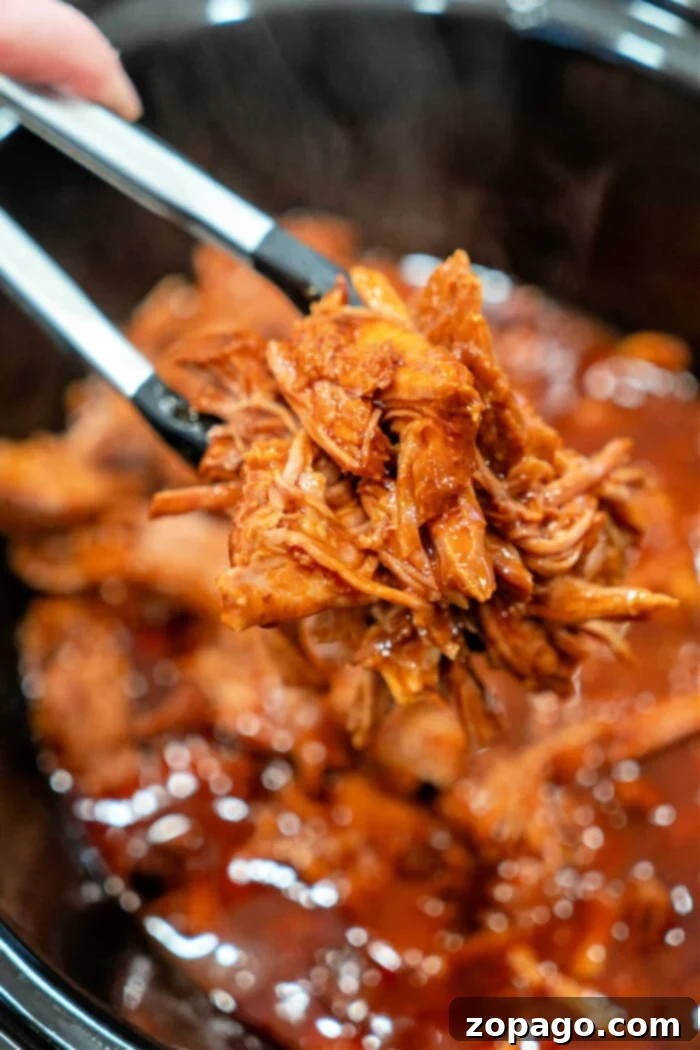 The Easiest 2-Ingredient Pulled Pork 2 Tongs holding shredded BBQ pulled pork over a slow cooker full of this mouthwatering 2 ingredient pulled pork recipe.
