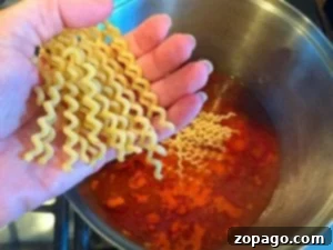 Streamlined Spaghetti Supper 7 A hand gracefully holds uncooked wavy pasta over a pot of vibrant orange-hued liquid simmering on the stove, perfect for creating a delicious one-pot spaghetti masterpiece.