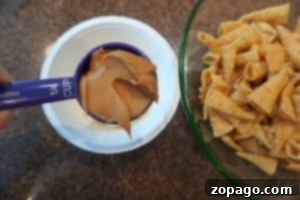A measuring cup filled with peanut butter is held above a white container, ready to create some unicorn horn muddy buddies. Next to it, a green bowl holds a pile of rolled corn snacks, resting on the brown countertop in perfect harmony.