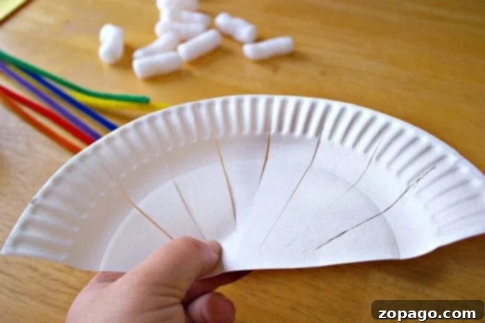Kaleidoscope Weaving 3 A child's hand holds a pre-cut white paper plate with multiple slits ready for rainbow weaving. Scattered colorful pipe cleaners and white packing peanuts lie on a wooden background, setting a playful craft scene.