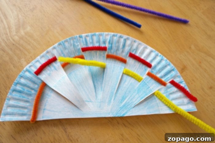 Kaleidoscope Weaving 4 A bright rainbow weaving art craft in progress, featuring a blue paper plate fan-shaped base with red, yellow, and orange pipe cleaners woven through the slits, resting on a wooden table.