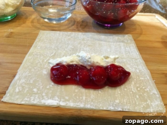 Cherry cheesecake egg rolls are amazing!! Just 3 ingredients in this warm dessert egg roll recipe. SO easy and the easiest cheesecake recipe ever. #cherrycheesecake #cheesecake #eggrolls #desserteggrolls #cherry #thetypicalmom Square pastry sheet topped with cherry pie filling and cream cheese on a wooden board, reminiscent of cherry cheesecake egg rolls, with a bowl of cherry filling and water in the background.