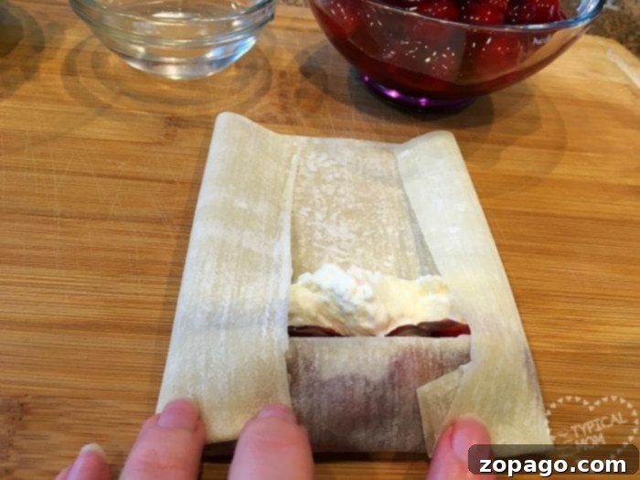 Cherry cheesecake egg rolls are amazing!! Just 3 ingredients in this warm dessert egg roll recipe. SO easy and the easiest cheesecake recipe ever. #cherrycheesecake #cheesecake #eggrolls #desserteggrolls #cherry #thetypicalmom A person skillfully folds a egg roll wrapper filled with creamy cherry cheesecake and fruit filling on a wooden board.