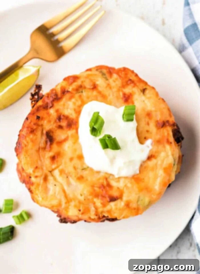 Crispy Cheesy Potato Cakes 2 Crispy potato cake topped with sour cream and green onions on a white plate, alongside a lime wedge, fork, and an enticing dollop of fried mashed potatoes with cheese.