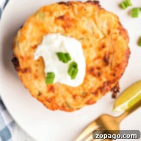 A cheesy potato pancake topped with sour cream and chopped green onions, served with lime wedges on a white plate, reminiscent of fried mashed potatoes with cheese.