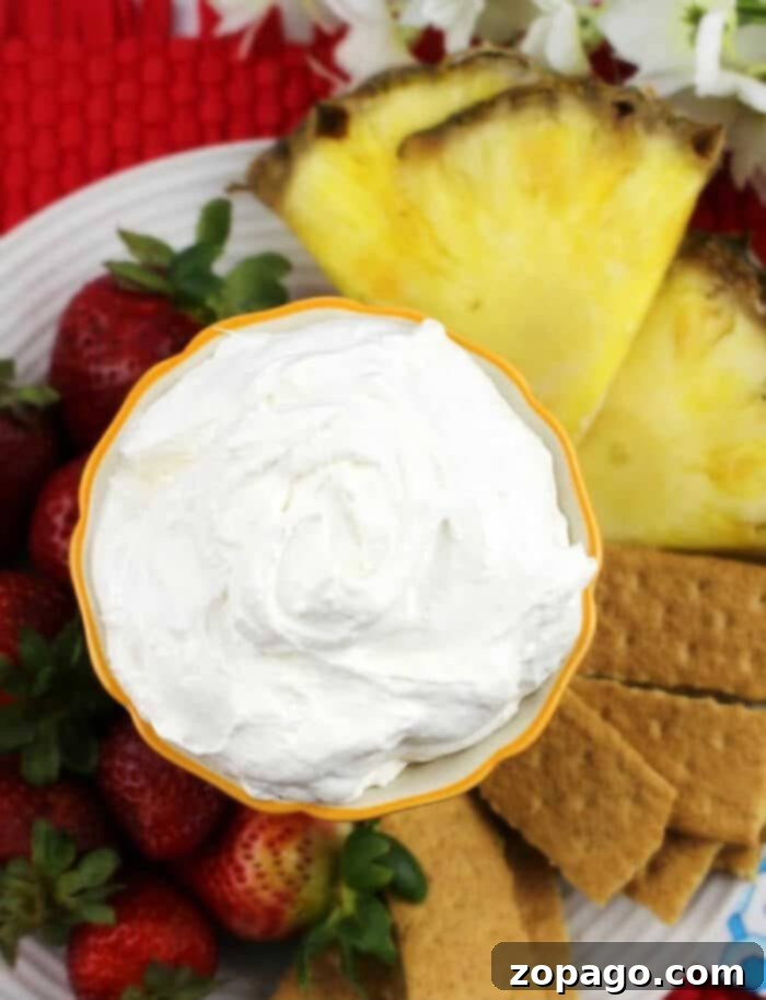 Creamy Pineapple Delight Dip 6 A top-down view of a large, clear glass bowl filled with creamy Pina Colada dip, surrounded by an abundance of fresh, ripe pineapple slices, vibrant red strawberries, and crunchy graham cracker squares. The arrangement is festive and inviting, suggesting a perfect dessert or appetizer for a summer party, showcasing the versatility of this tropical dip with various dippers.