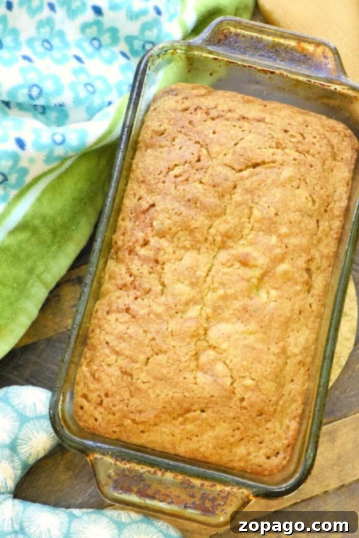 A freshly baked loaf of Zucchini Pineapple Bread, still in its glass baking pan, resting on a wooden surface with beautifully patterned towels nearby, highlighting its golden crust and inviting texture.