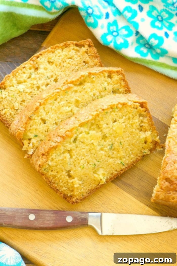 Beautifully sliced Zucchini Pineapple Bread arranged on a wooden cutting board, with a knife alongside. A green and blue floral towel provides a rustic backdrop, showcasing the bread's moist texture and golden crust.