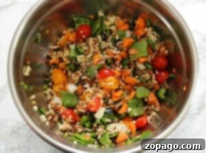A metal bowl filled with mixed vegetables, cherry tomatoes, rice, and slices of grilled stuffed peppers sits on a light marble surface.