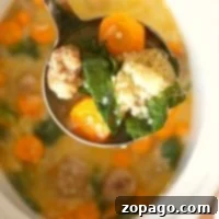A ladle of easy Italian wedding soup with meatballs, carrots, spinach, and couscous hovers over a bowl.