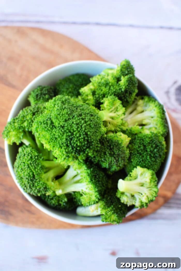 Crisp-Tender Oven Broccoli 2 A bowl filled with fresh green broccoli florets, perfectly prepared for a delicious steamed broccoli recipe, is placed on a rustic wooden cutting board. The background shows a lightly textured white surface, highlighting the vibrant green of the vegetable.
