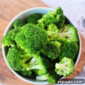 A bowl filled with fresh green broccoli florets, perfect for a delicious steamed broccoli recipe, is placed on a wooden cutting board. The background shows a lightly textured white surface.
