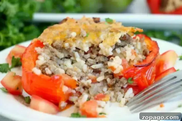 Effortless Bell Pepper Blanching 6 A stuffed red bell pepper, blanched to perfection, sits on a white plate filled with a savory mix of ground meat and rice, topped with melted cheese. Diced tomatoes and chopped herbs are artfully scattered around the pepper, while a fork rests beside it.