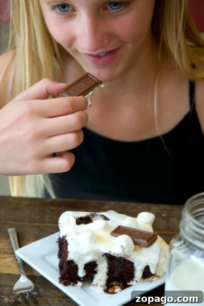 This S'mores Poke Cake is a dream for chocolate and marshmallow enthusiasts! A perfect, easy-to-make dessert for enjoying classic s'mores flavors at home. Features rich chocolate cake soaked in chocolate, topped generously with marshmallows and graham crackers. #pokecakerecipe #smoresdessert #chocolateandmarshmallow #easybaking #dessertideas Close-up of a S'mores Poke Cake slice showing layers of chocolate cake, sauce, and marshmallow topping
