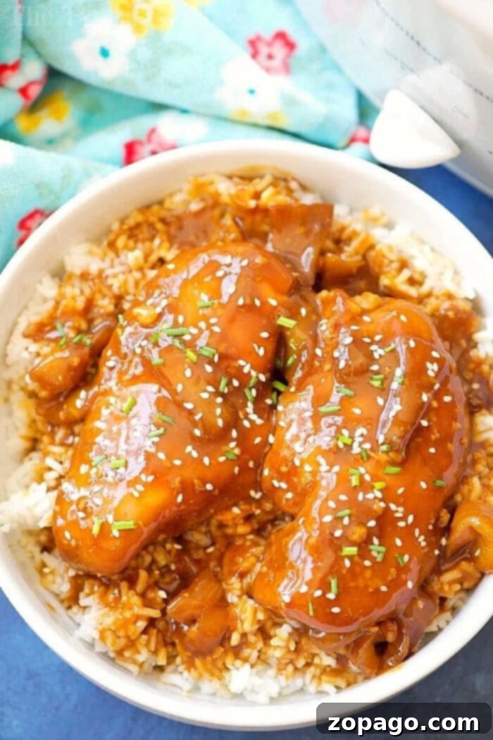 This easy Crockpot sticky teriyaki chicken with a kick of heat recipe is amazing and a healthy meal too! Sweet with just a pinch of Sriracha makes it great! Bowl of rice topped with Crockpot teriyaki chicken, chopped herbs, sesame seeds, and a savory brown sauce.