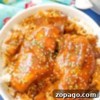 A bowl of rice topped with Crockpot teriyaki chicken pieces is garnished with sesame seeds and chopped greens. The dish is served on a blue surface with a floral-patterned cloth nearby.