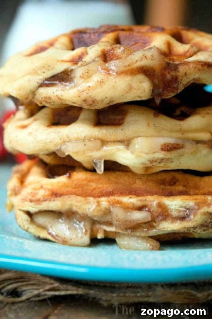 Cinnamon Roll Waffles 5 Close-up of a stack of three glazed, apple pie stuffed waffles on a blue plate. The waffles show a golden-brown, slightly crispy texture with a dripping glaze. The background is blurred, putting focus on the cinnamon roll waffles. Text "The Typical Mom" is faintly visible at the bottom right.