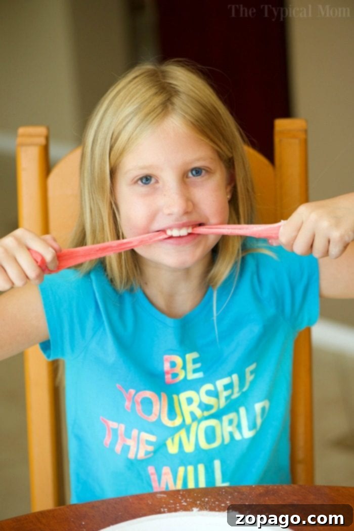 Fluffy Shaving Cream Slime 2 A child in a blue shirt stretches pink slime while sitting at a table, pondering how to make edible slime for an exciting twist.