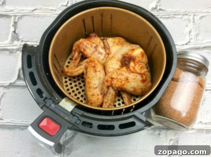 Here's an easy air fryer wings recipe and the best rub for chicken wings ever! If you're looking for a healthy alternative to fried chicken with the same great taste these naked chicken wings in your air fryer are the answer. Simple and flavorful without using any oil to cook them. #airfryer #chicken #wings #chickenwings #rub #best #naked #recipe #simple An air fryer wings recipe comes to life as a basket of perfectly seasoned chicken wings sits on a white brick background, accompanied by a jar filled with flavorful seasoning mix.