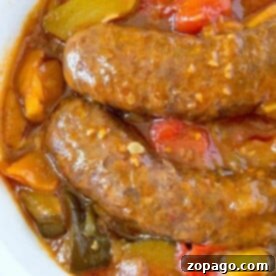Easy Instant Pot Sausage and Peppers 2 A white bowl contains two pressure cooker sausages submerged in a thick, chunky sauce with visible pieces of red, green, and yellow bell peppers. The sauce has a glossy texture, suggesting a rich, flavorful broth or gravy. The dish is set on a surface with a hint of colorful fabric.