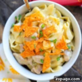 A bowl of creamy noodle casserole with chicken and crushed Doritos, served with a spoon and garnished with chives.