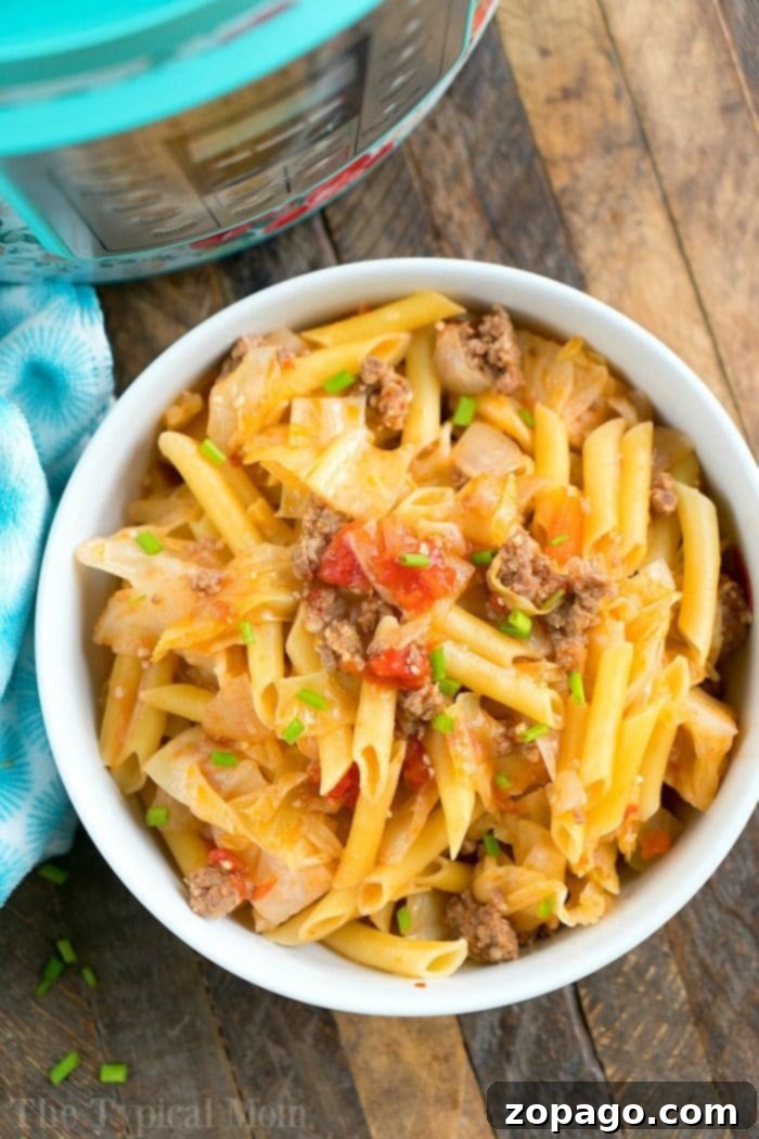 Delicious pressure cooker sausage and cabbage with noodles, a simple dinner idea packed with flavor. A bowl filled with penne pasta, perfectly cooked in a pressure cooker, mixed with ground beef, chopped tomatoes, and savory sausage. The dish is topped with green onions and accompanied by crisp cabbage on a wooden table beside a blue patterned cloth.