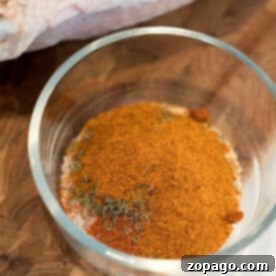 A glass bowl filled with a mix of dry spices for a smoked turkey rub recipe sits on a wooden surface next to raw poultry.