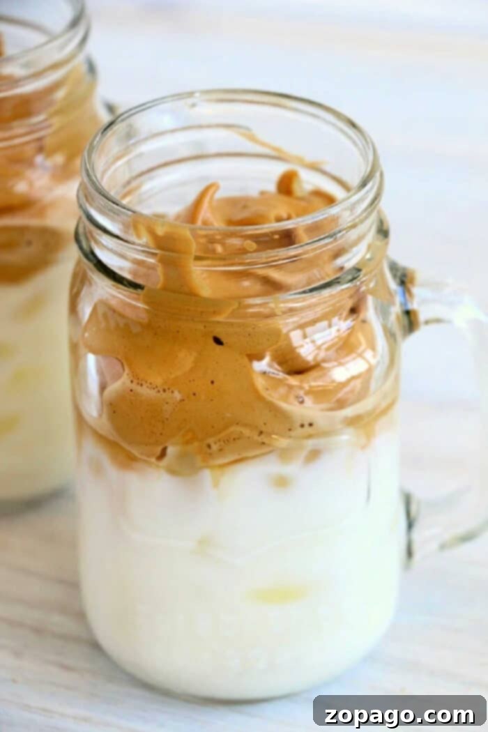 Try this 3 ingredient trendy Dalgona whipped coffee recipe! EASY to make this popular TikTok drink everyone is going crazy over. I bet you'll love it too. A beautifully layered Dalgona whipped coffee, featuring a thick, frothy coffee foam topping a glass of milk with ice cubes. Garnished with a light dusting of cocoa powder.