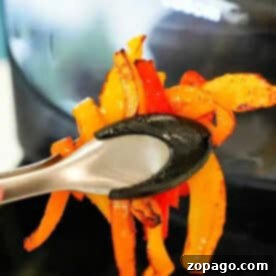 Tongs holding cooked orange and red air fryer peppers in front of an air fryer.