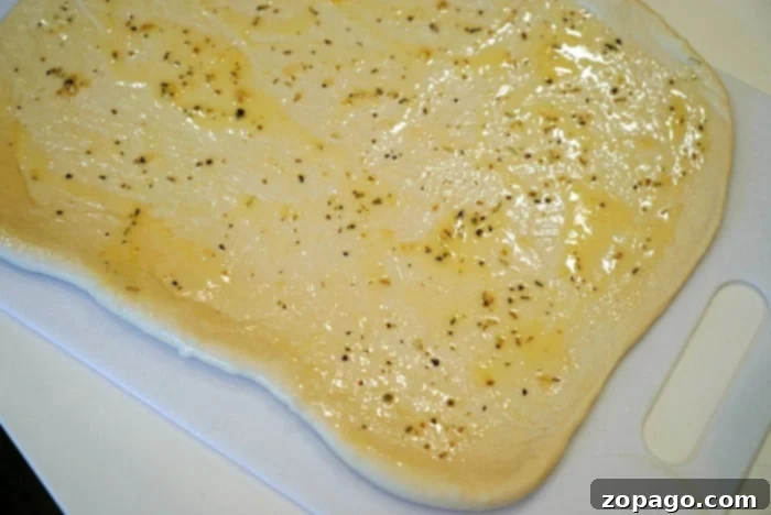 Pizza Swirls 4 A sheet of uncooked thawed from frozen pizza dough is evenly spread with olive oil and sprinkled with herbs, positioned on a white cutting board, ready for toppings.