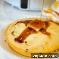 Air Fryer Pancake Perfection 8 An air fryer pancake made with Bisquick is topped with a pat of butter as syrup is drizzled over it. The golden delight rests on a white plate, with a floral-patterned cloth partially visible in the background, adding a charming touch to this breakfast scene.