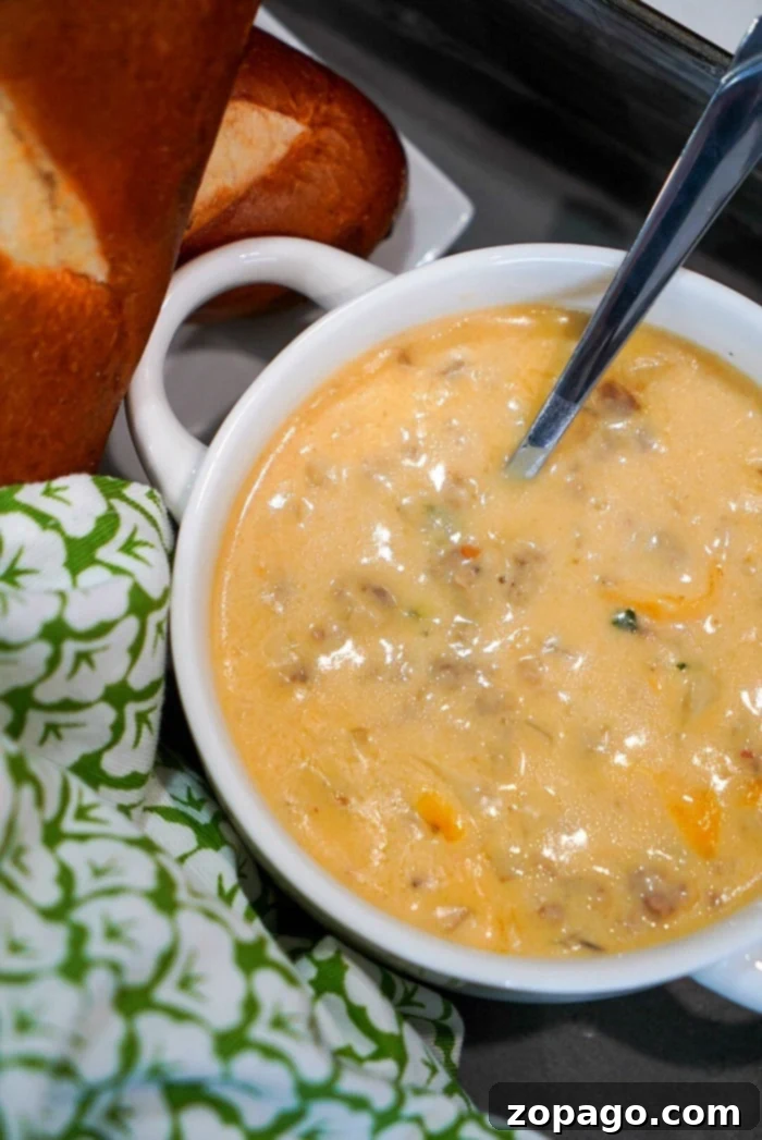 The Ultimate Instant Pot Beer Cheese Soup 2 A bowl of creamy beer cheese soup with visible chunks, a spoon inside, bread, and a green patterned cloth nearby evokes the comforting scent of an Instant Pot creation.
