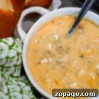 The Ultimate Instant Pot Beer Cheese Soup 5 A bowl of creamy beer cheese soup with visible chunks, a spoon inside, bread, and a green patterned cloth nearby evokes the comforting scent of an Instant Pot creation.