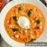A white bowl brimming with creamy instant pot buffalo chicken soup is garnished with a dollop of sour cream. This savory delight includes chunks of vegetables and herbs, vibrant red and yellow peppers, and bits of blue cheese scattered on top.