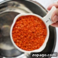 Easy Instant Pot Red Lentils 6 A hand holds a 1-cup measuring cup brimming with vibrant red lentils over a metal bowl, ready for the instant pot.