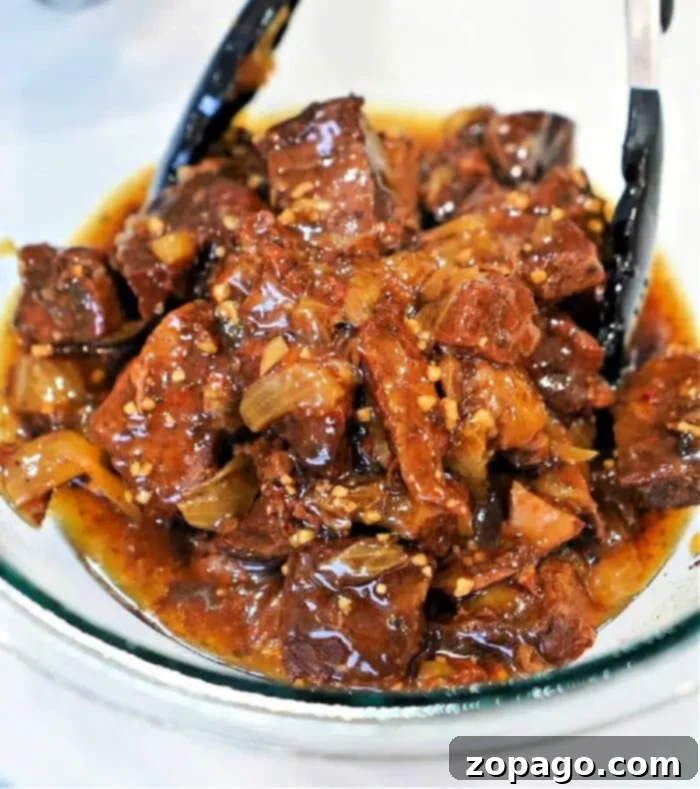 Perfect Beef Arm Roast 11 A glass bowl filled with beef stew, showcasing a classic beef arm roast recipe with chunks of beef and onions in a rich, dark sauce. Black tongs rest in the bowl, and the stew is visibly seasoned with fresh herbs and spices.