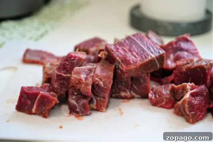 Perfect Beef Arm Roast 5 Chopped pieces of raw beef arm roast pieces are placed on a white cutting board, perfect for starting a beef arm roast recipe. The meat is marbled with fat, and the board rests on a kitchen counter. The background features a green-patterned cloth and part of a round, gray object.